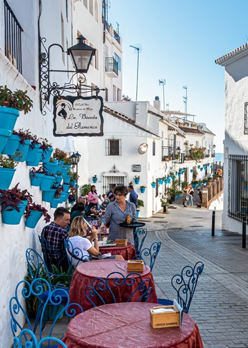 A cafe serving drinks to customers on a street full of blue plant pots and white buildings