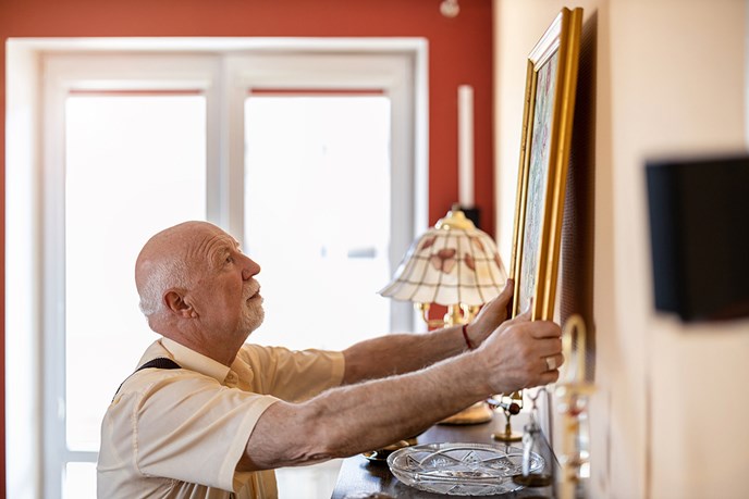 An elderly man putting up a painting on a wall in his home.